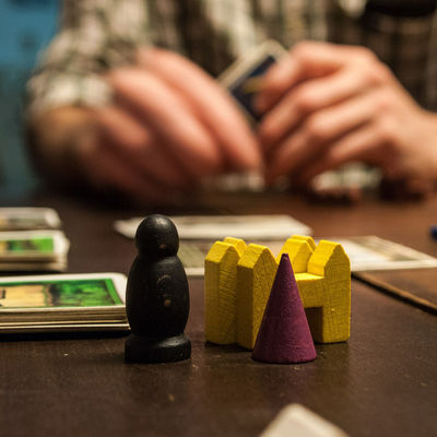 auf einem Tisch liegen karten und stehen Spielsteine aus Holz. Im Hintergrund sitzt jemand und hlt Karten in der Hand
