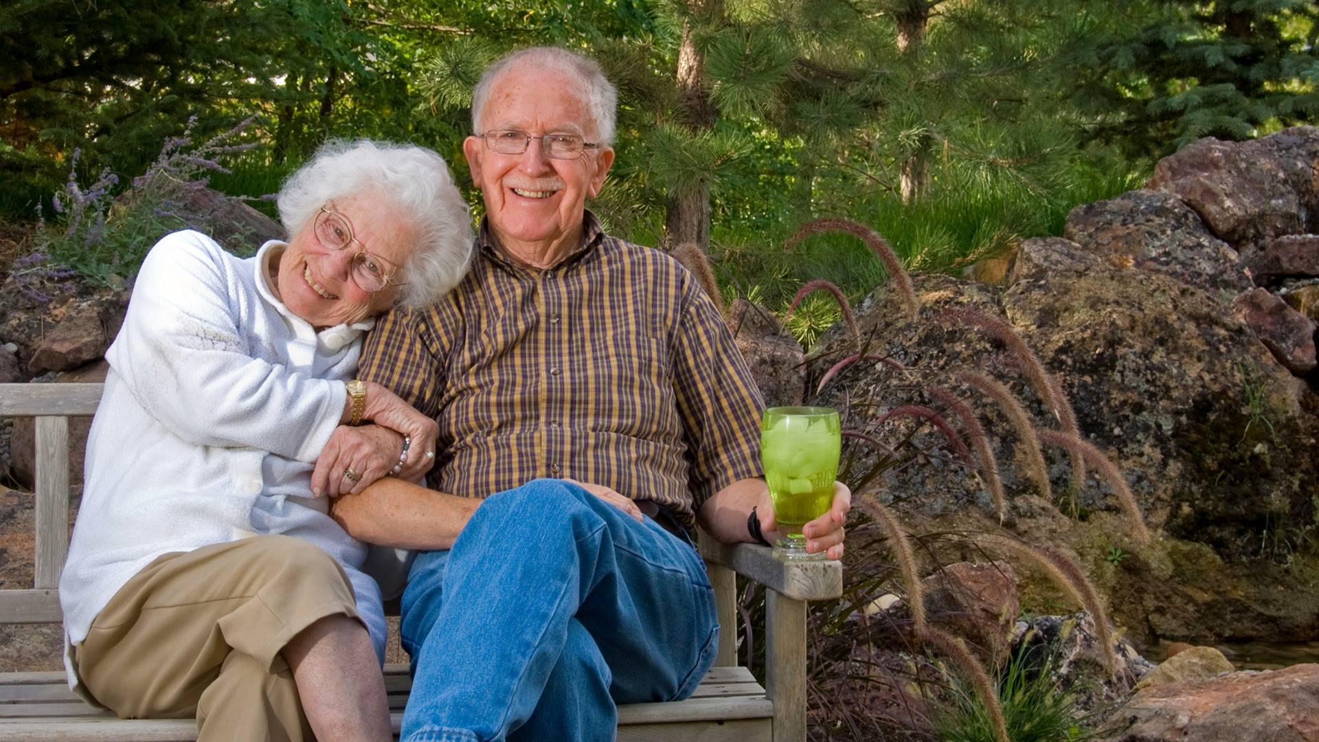Ein Seniorenpärchen sitzt auf einer Bank. Die Frau hält sich am Arm des Mannes fest. Beide lachen. Der Mann hält ein gefülltes Glas in der Hand.