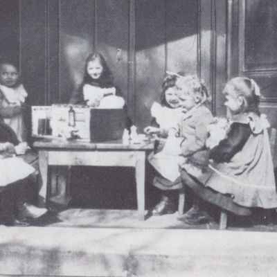 historisches Schwarzweißfoto von sieben Mädchen des Henriette Breymann Kindergarten, die an einem Kindertisch sitzen oder stehen.