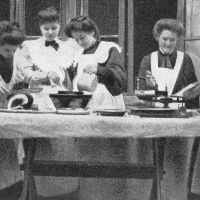 historisches schwarzweiß-Foto: fünf frauen mit weißen Rüschenschürzen stehen an einem Tisch und scheinen zu backen