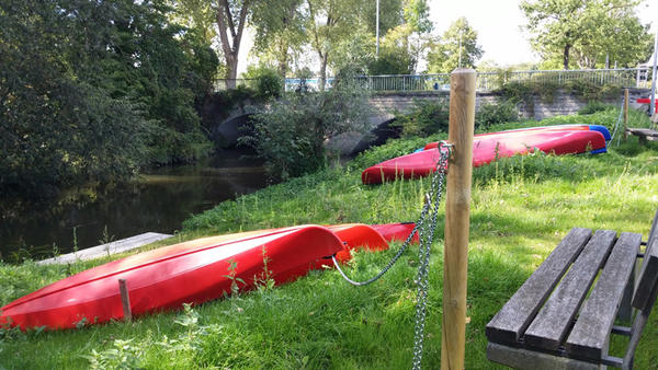 An einem Flussufer neben einer Bank liegen mehrere Kanus auf dem Rasen.