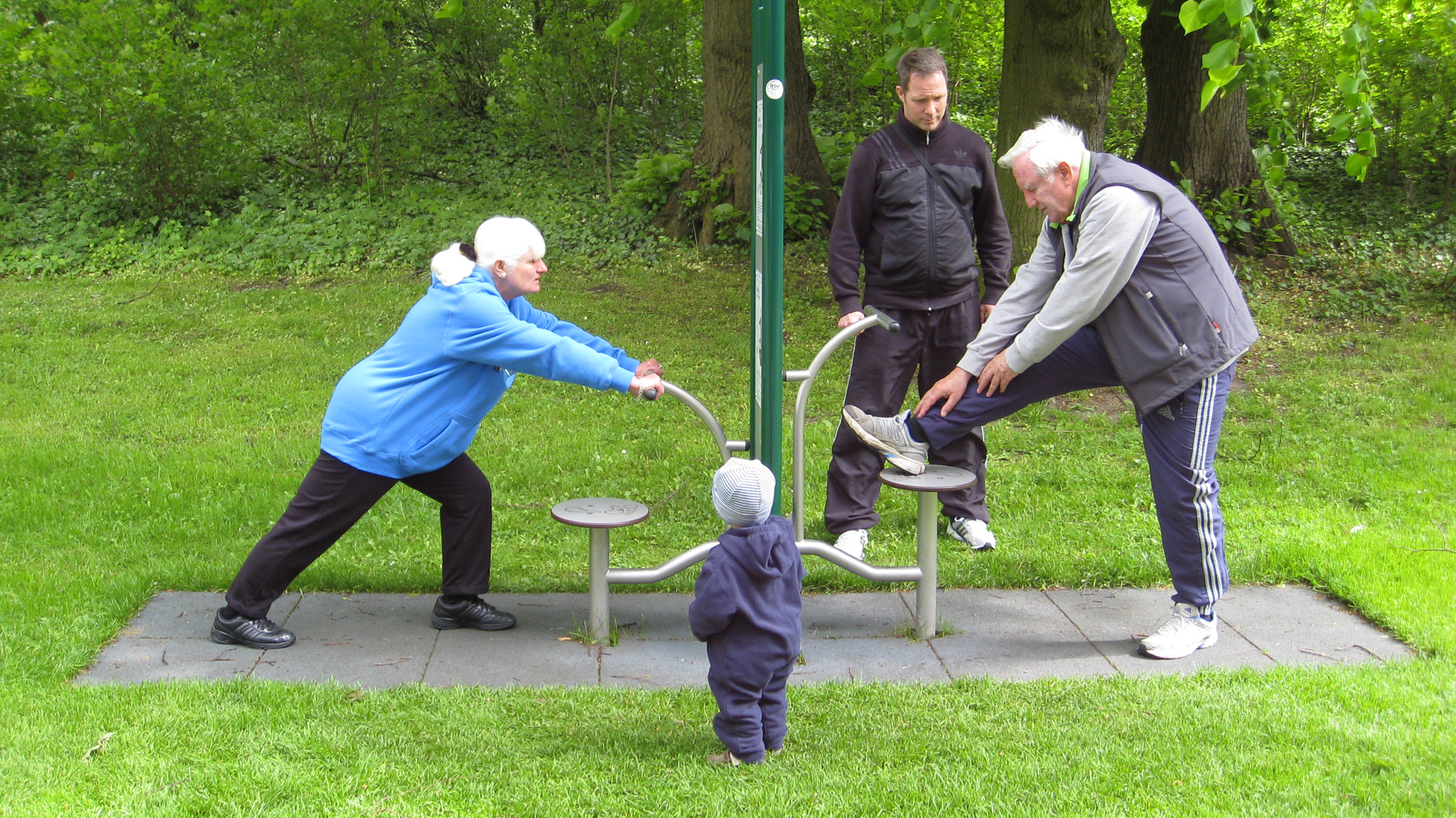 An einem Outdoor-Fitnessgerät in einer Rasenfläche trainieren eine ältere Frau und ein älterer mann. Ein anderer Mann im Trainingsanzug schaut zu, ebenso ein kleines Kind