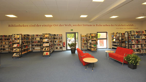 An einer langen Wand in der Stadtbücherei stehen Bücherregale, davor zwei rote Sofas mit Tischchen und Grünpflanzen.