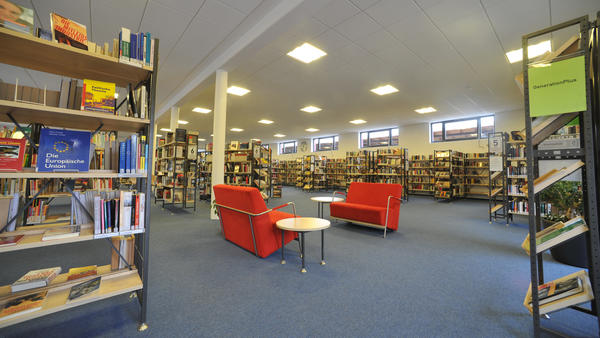 In einem großen Raum stehen viele Bücherregale, in der Raummitte zwei rote Sofas mit Tischchen.
