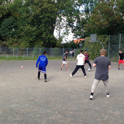 Jugnen spielen auf einem Aschenplatz Fußball. An der Seite an einem Zaun hängt ein Basketballkorb