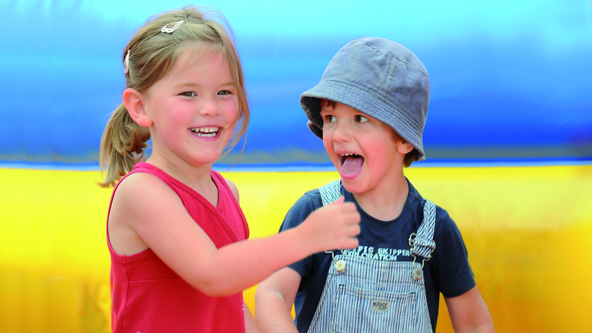 Vor einem blau-gelben Hintergrund stehen links ein kleines Mädchen und rechts ein kleiner Junge mit Anglerhut nebeneinander.