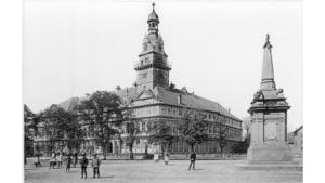 Auf einem großen Platz stehen mehrere Personen und rechts im Bild steht ein Monument auf dem Platz. Im Hintergrund steht ein großes Gebäude mit Turm und davor einige Bäume.