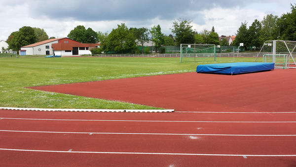 Neben einer grünen Graswiese sind im Vordergrund rote Flächen zu sehen. Im Hintergrund des Sportplatzes mit der roten Laufbahn steht ein Gebäude.