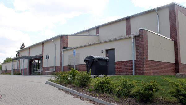 Fassadenansicht eines großen Gebäudes. Vor dem Gebäude wachsen grüne Sträucher und im Mittelpunkt steht eine schwarze große Mülltonne.
