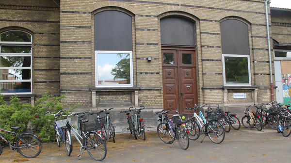 Fassade eines Gebäudes vor dem einige Fahrräder stehen. Einige Fenster und eine doppelflügelige braune Eingangstür sind zu erkennen.