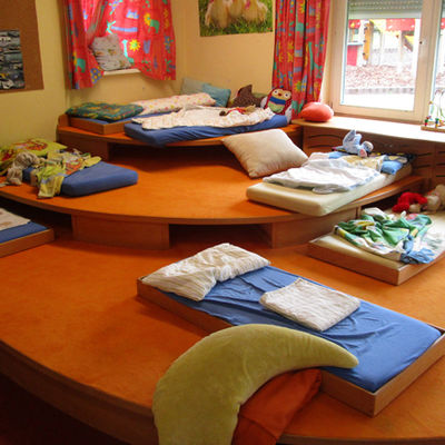 In einem Innenraum mit drei aufeinander aufgebauten Holzpodesten sind mehrere Matratzen mit Kissen und Decken platziert.