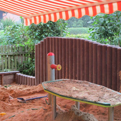 Vor einem Holzpalisaden-Zaun steht ein Tisch auf dem Sand liegt. Auch auf dem Boden liegt Sand und kleinere Kunststoffschaufeln. Überdacht ist die Szenerie von einer ausgefahrenen rot-weißen Markise.