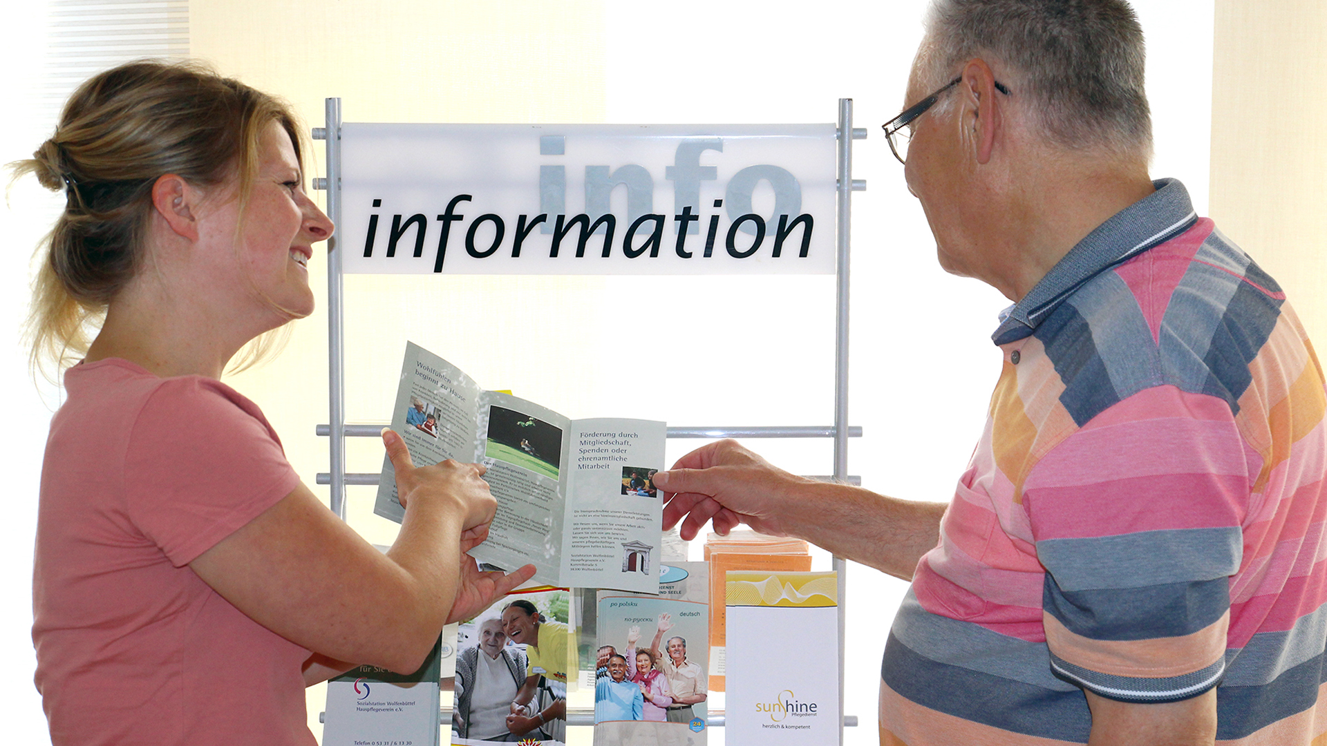 Eine junge Frau steht mit einem älteren Herrn an einem Prospektständer mit der Aufschrift "Information" und zeigt ihm einen aufgeschlagenen Informationsflyer.