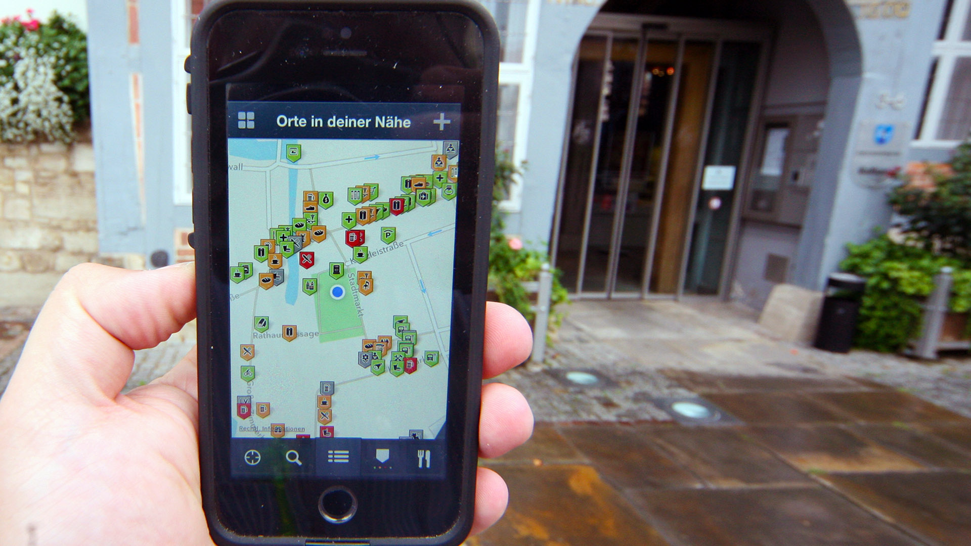 Eine Hand hält ein Smartphone mit der geöffneten Wheelmap-App hoch. Rechts daneben ist der Eingang des Rathauses zu sehen.