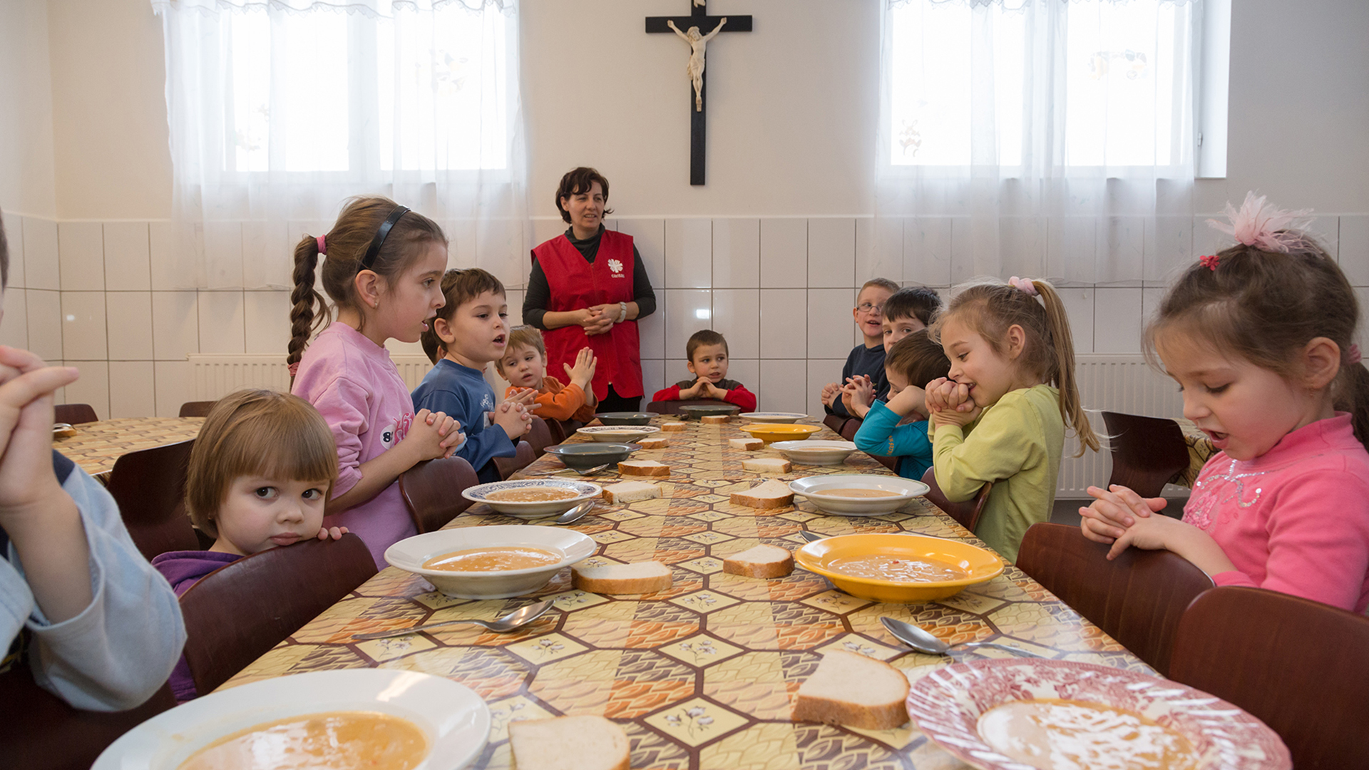 Gruppenfoto von Kindern und einer Frau an einem gemeinsamen Essenstisch mit Suppentellern. Im Hintergrund ist ein Holzkreuz an einer Wand zu erkennen.