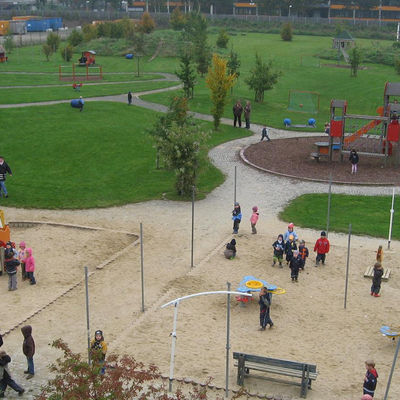 Luftaufnahme eines Spielplatzes mit Sand- und Grünflächen, auf dem Kinder spielen.