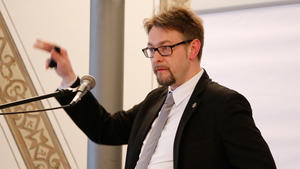 Ein Mann mit Anzug und Brille gekleidet steht vor einem Mikrofon und macht eine Handbewegung.