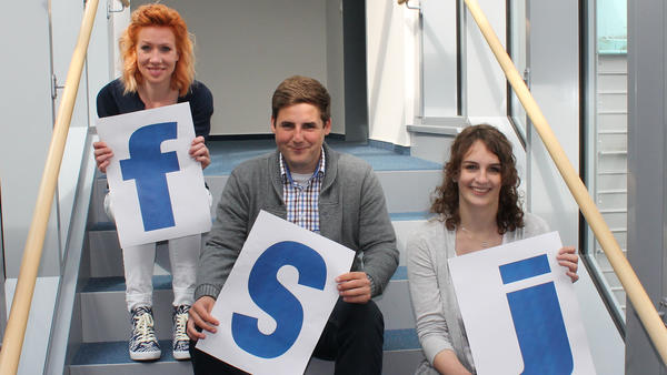Drei junge Menschen sitzen auf Treppenstufen und halten je einen Buchstaben in der Hand, die zusammen das Wort FSJ ergeben.