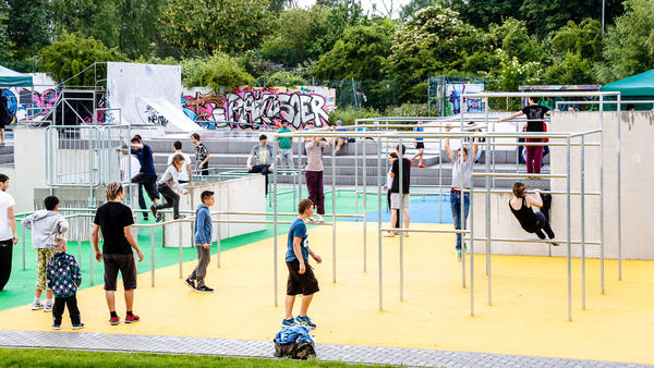 Kinder und Jugendliche spielen auf dem Jugendplatz an Klettergerüsten auf der Parkour-Anlage, im Hintergrund sieht man Beton-Sitzstufen und den Skaterpark.