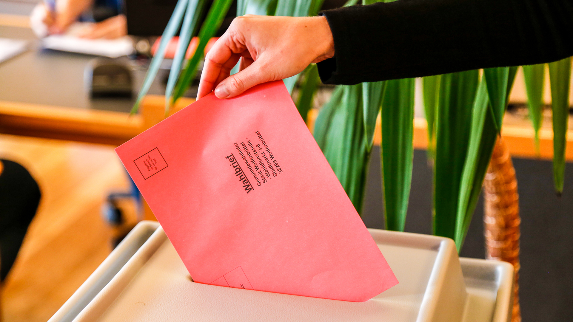 Ein Wahlbrief mit rotem Umschlag wird von einer Hand durch einen oberseitigen Schlitz in eine Wahlurne eingeworfen.
