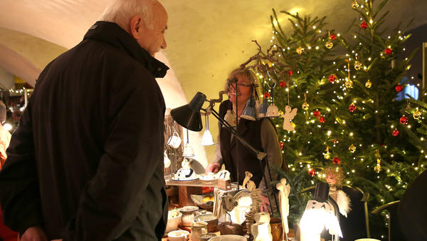 In einem Kellergewölbe steht eine Frau hinter einem kleinen Verkaufsstand. Vor ihr schaut sich ein Mann die ausgelegten Gegenstände an. Hinter dieser Szenerie sind mit Lichterketten geschmückte Weihnachtsbäume aufgestellt.