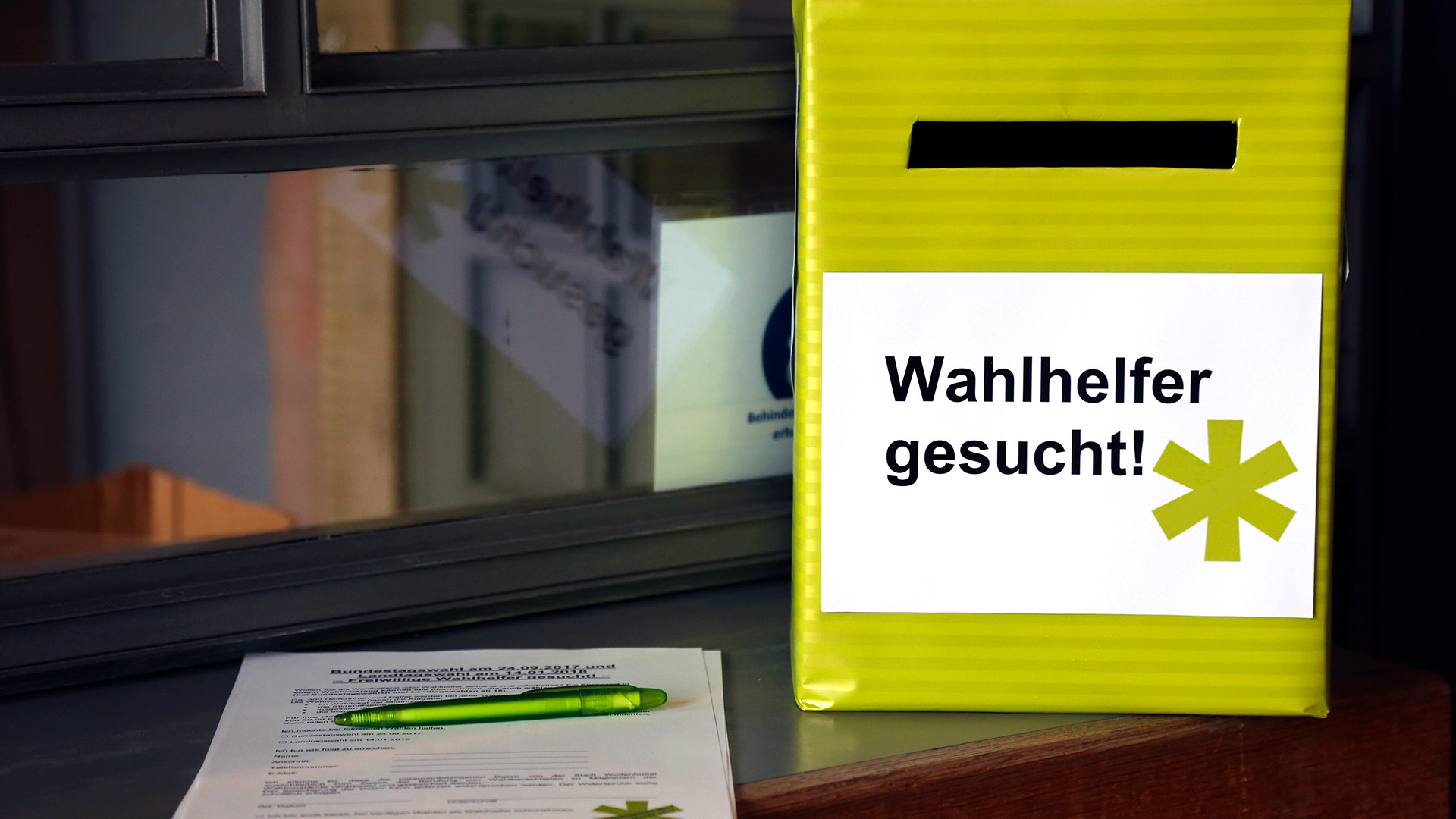 Auf einer grünen Einwurfbox mit Einwurfschlitz steht "Wahlhelfer gesucht". Daneben liegen Anmeldebögen und ein Stift.