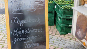 Auf einem Schild an einem Verkaufsstand auf einem Wochenmarkt steht: "Tutscheks. Doppelback. Mohnstriezel. Gebackene Torten."