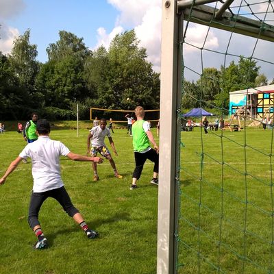 Auf einer grünen Wiese spielt eine junge Menschen Fußball. Außerdem ist ein Tor zu sehen, in dem ein Torhüter mit Torwart-Handschuhen steht.