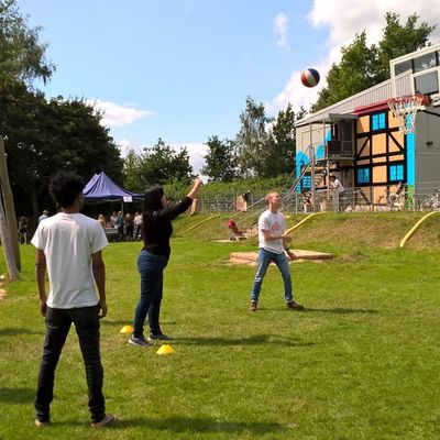 Jugendliche werfen mit einem Ball auf einen Basketballkorb. Das Spiel findet auf einer Wiese statt.