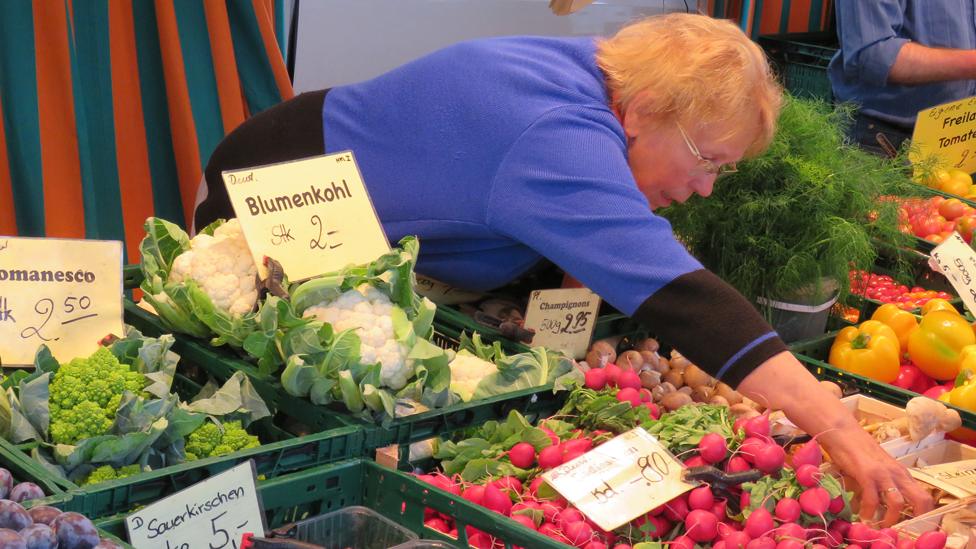 Eine Frau eines Verkaufsstandes auf einem Markt beugt sich über Kisten verschiedener Gemüsesorten.