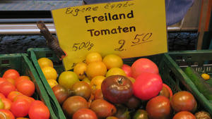 Im Mittelpunkt steht eine grüne Kiste eines Verkaufsstandes auf einem Markt, die verschiedenfarbige Tomaten enthält. An die Kiste ist mit einer Klammer eine gelbes Schild geheftet und besagt: "Eigene Ernte. Freiland Tomaten. 500 Gramm. 2,50 Euro.