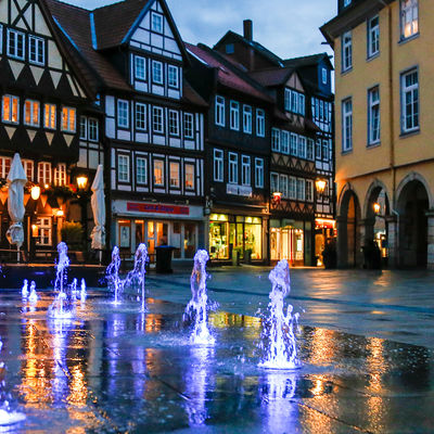 Brunnen in der Innenstadt Wolfenbüttels mit Beleuchtung in der Abenddämmerung. Im _Hintergrund sind Fachwerk-Gebäude zu erkennen.