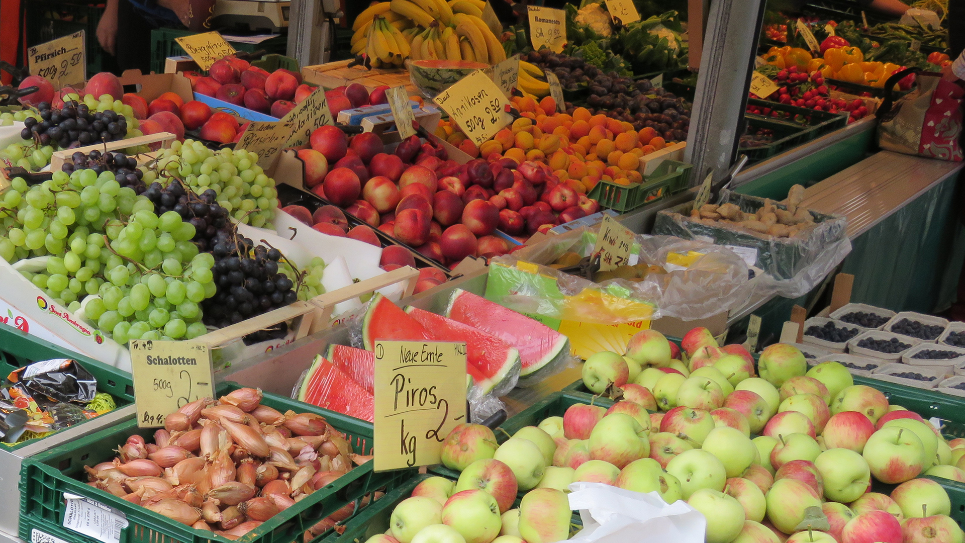 An einem Verkaufsstand werden im Vordergrund verschiedene Obstsorten angeboten.