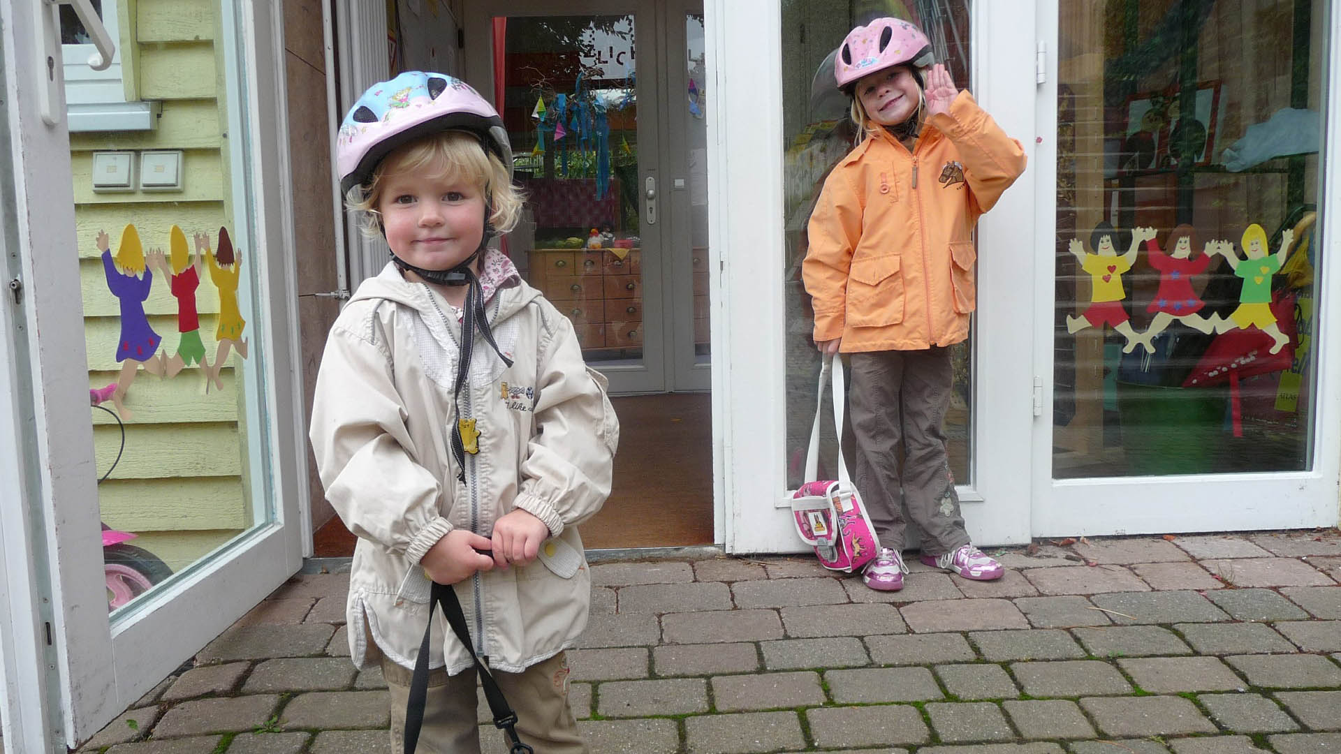 Vor dem Eingangsbereich eines Kindergartens stehen zwei kleine Mädchen mit Fahrradhelmen gekleidet.