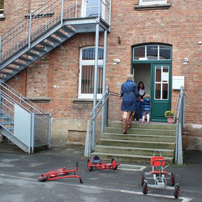 Ein Kind und zwei Erwachsene gehen in ein Gebäude. Vor der Treppe zum Eingang liegen Roller und Fahrzeuge für Kinder.