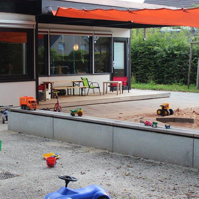Vor einer Kindertagesstätte liegt eine Sandplatz mit Sitzbänken, auf denen Kinderspielzeuge platziert sind.