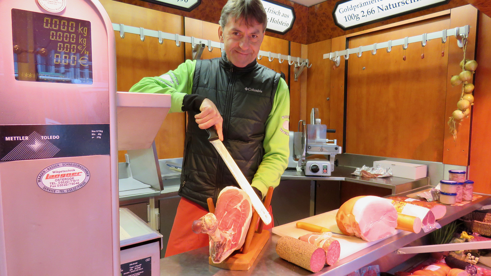 In einem Verkaufswagen auf dem Wolfenbütteler Wochenmarkt steht ein Mann hinter einem Verkaufstresen und schneidet mit einem Messer eine Scheibe Schinken.