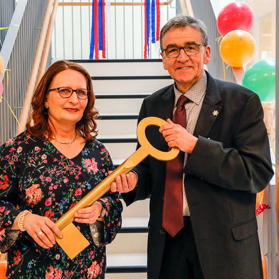 Vor einer Treppe stehen Bürgermeister Thomas Pink und die Leiterin der Kindertagesstätte Varierta Claudia Heesebeck mit einem großen Schlüssel zur symbolischen Schlüsselübergabe.