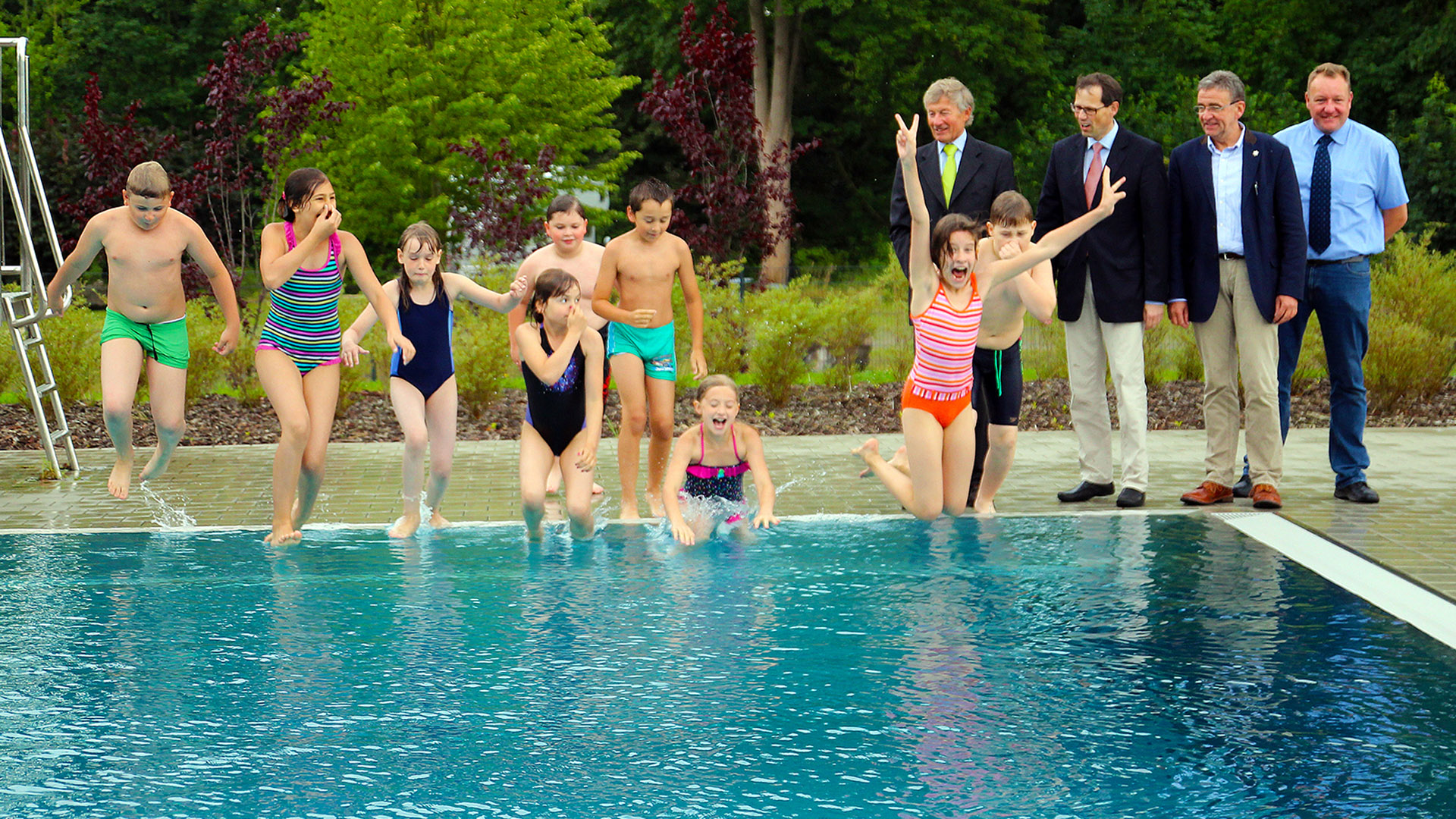 Kinder springen vom Beckenrand in ein Schwimmbecken. Daneben stehen Männer in Anzügen.