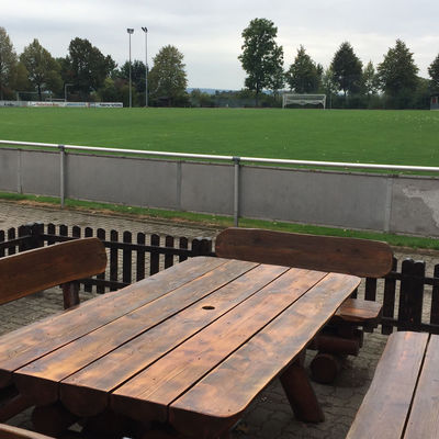Fußballplatz mit grünem Rasen und Tor. Im Vordergrund hinter den Sportplatz begrenzenden Banden steht eine hölzerne Sitzgruppe mit Bänken und einem Tisch.
