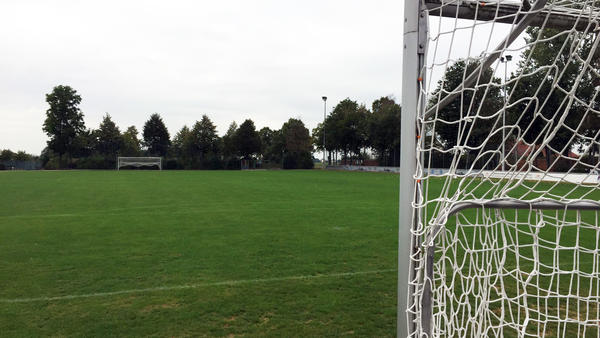 Hinter einem Fußballtor platziert, zeigt das Foto einen Fußballplatz. auf der gegenüberliegenden Seite steht ein zweites Tor mit weißem Netz. Der Platz ist stellenweise von höheren Bäumen umgeben.