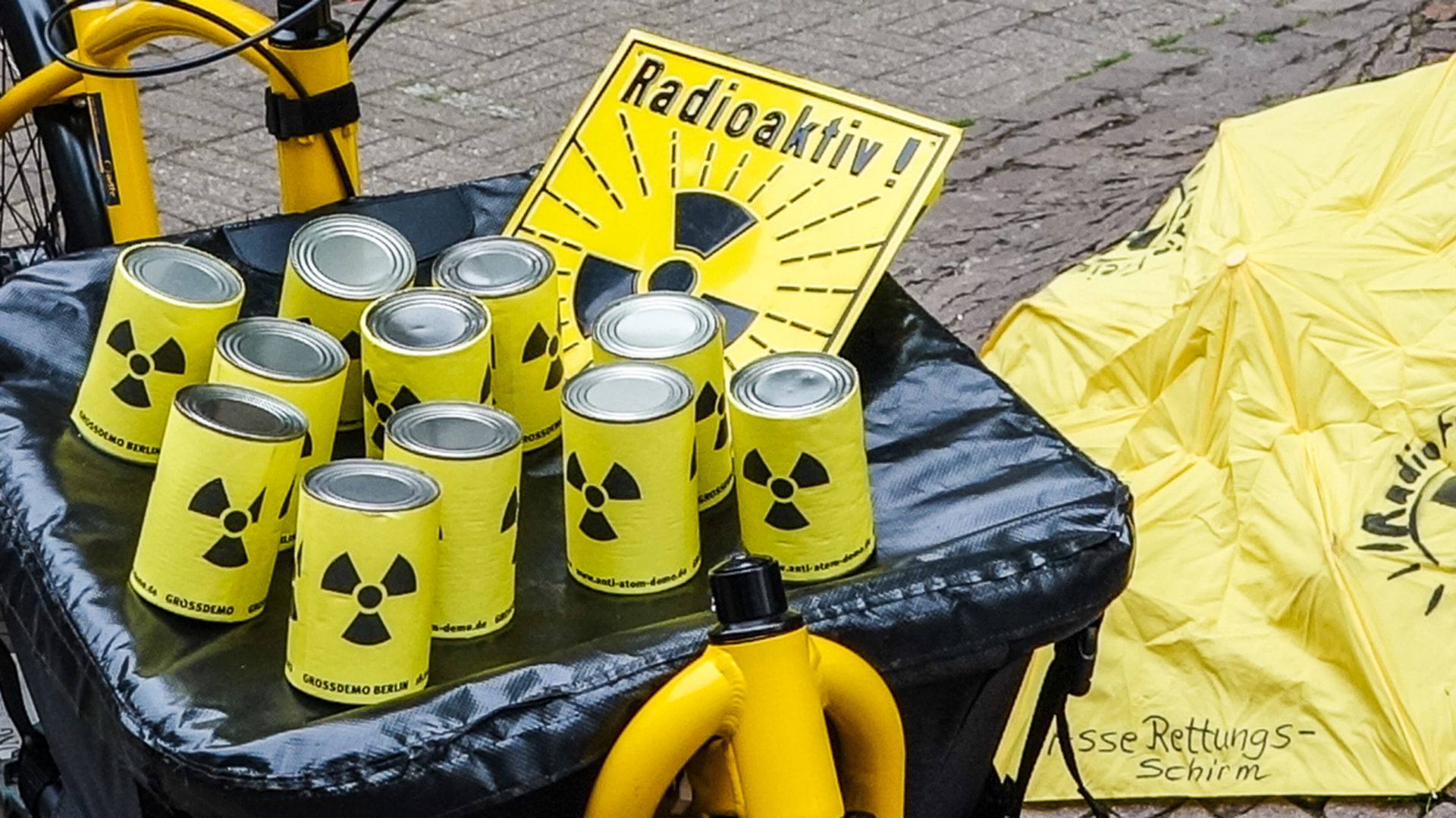 Auf dem Behälter eines Lastenrades stehen gelbe Dosen mit schwarzem Atom-Warn-Zeichen. Daneben liegt ein gelbes Schild mit gleichem Aufdruck wie die Dosen und dem Schriftzug "Radioaktiv!"
