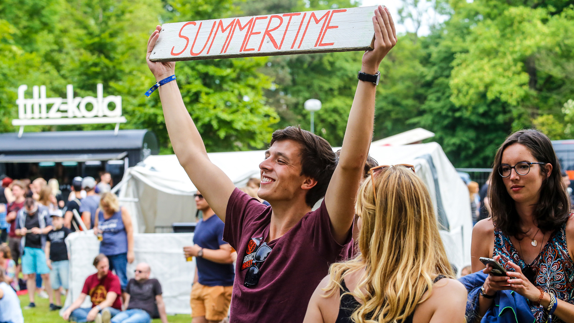 Ein junger Mann hält ein Schild mit der Aufschrift "Summertime" in die Luft. Direkt daneben stehen zwei jüngere Frauen.