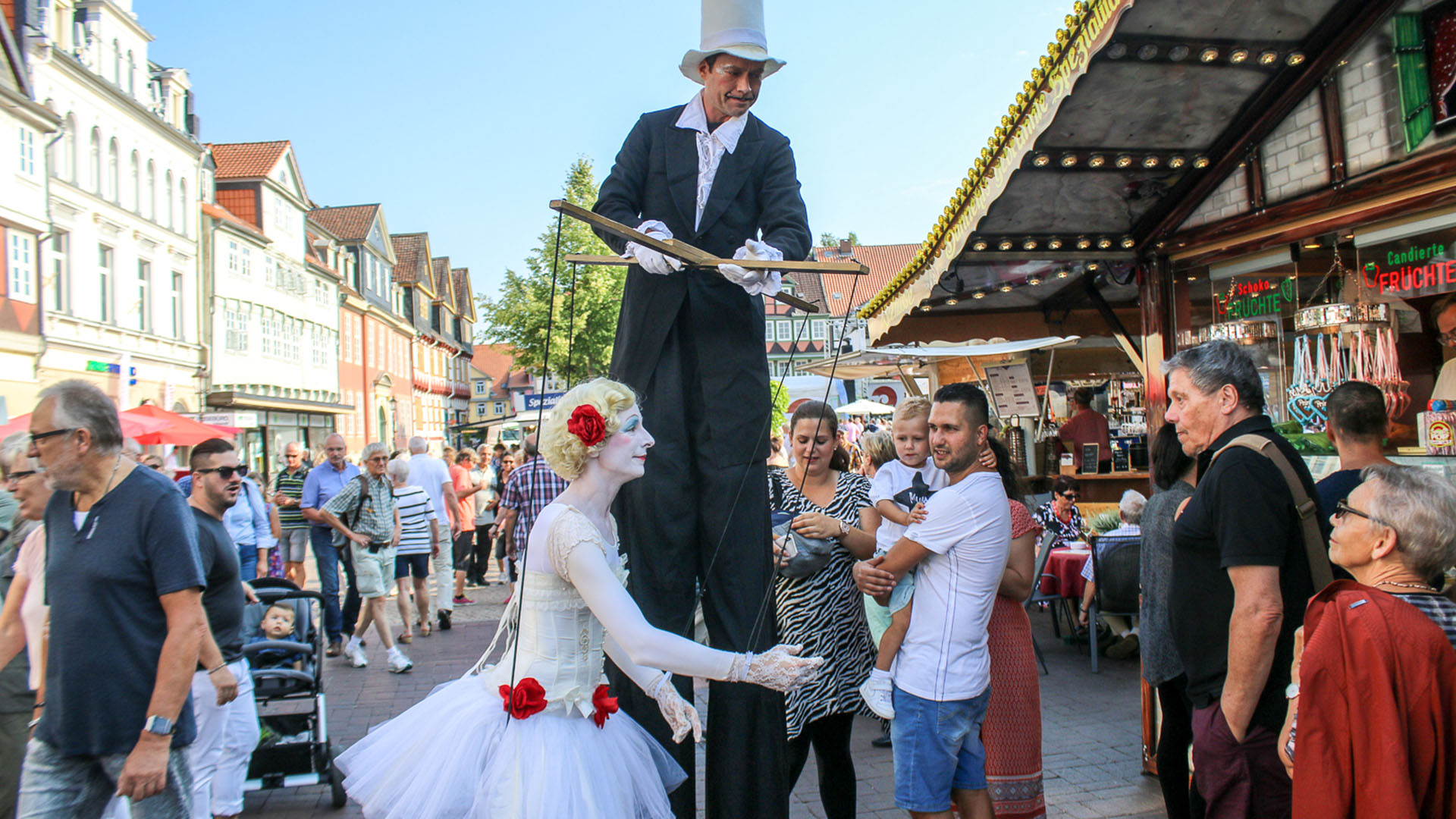 Ein Stelzenläufer wandert durch das Publikum in der Fußgängerzone und scheint eine Marionette zu bedienen, die in Realität eine Frau in Ballerina-Kostüm ist.