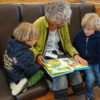 Eine Frau sitzt zwischen zwei Kindern. Alle drei Personen schauen in ein Kinderbuch, dass die Frau aufgeschlagen auf ihrem Schoß platziert hat.