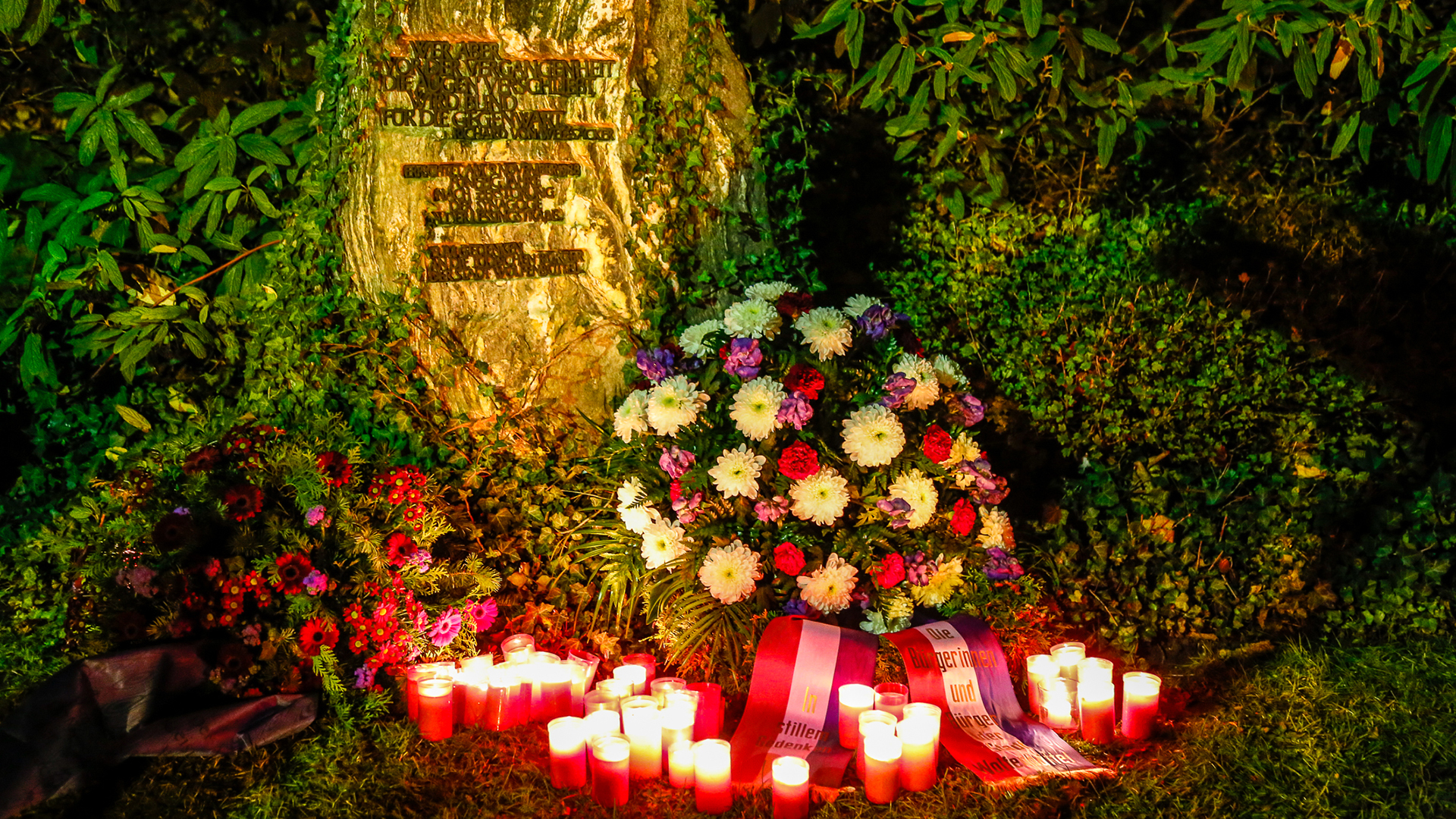 Gedenkstein mit Inschrift, einem niedergelegten Blumenkranz und mehreren angezündeten Grabkerzen.