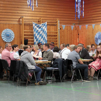 In einer Turnhalle sitzen Menschen an verschiedenen Tischreihen. An der Wand sind blau-weiße Fahnen angebracht.