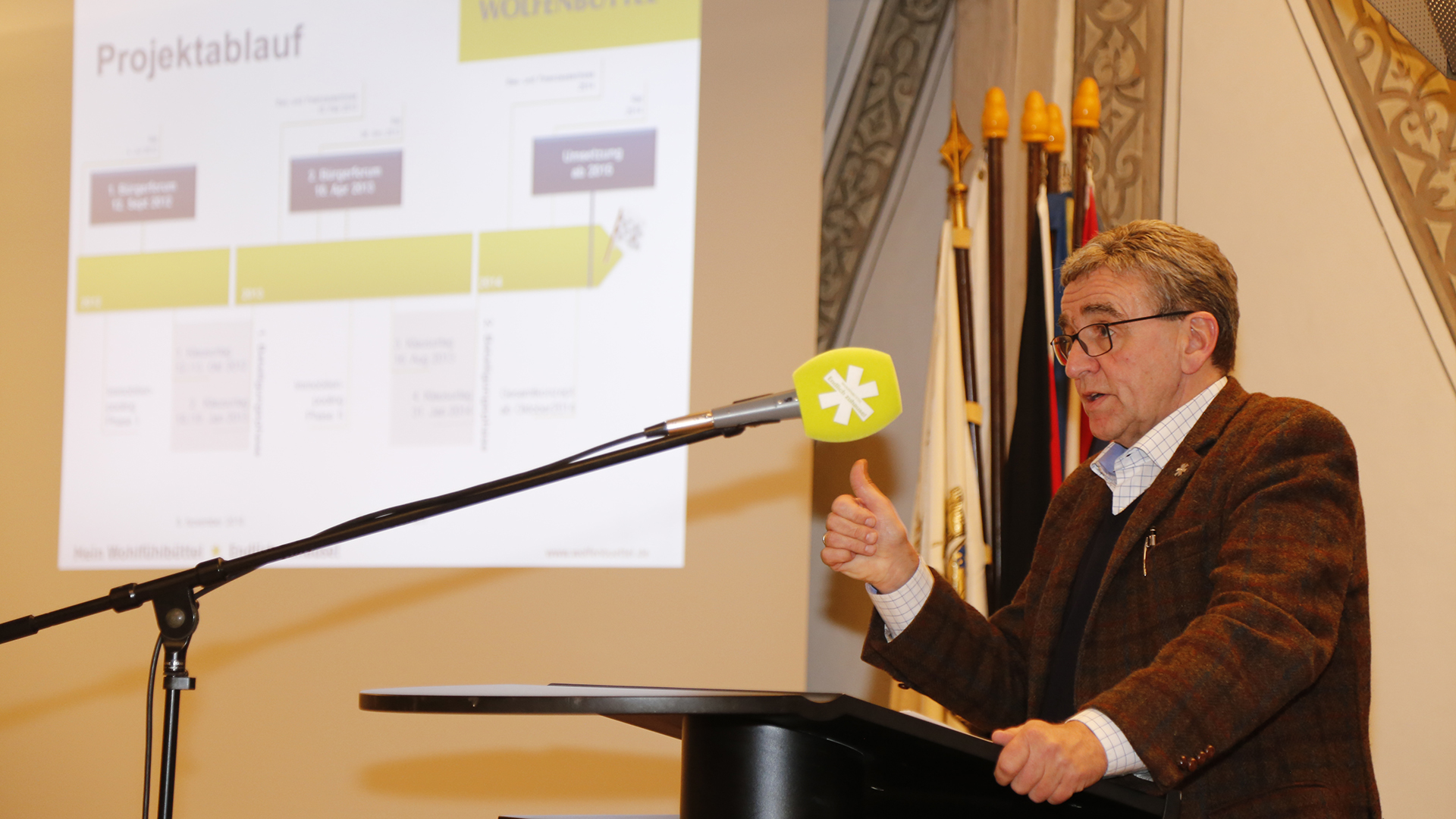 Thomas Pink spricht in ein grünes Mikrofon. Im Hintergrund eine Leinwand mit einer Grafik "Projektablauf".