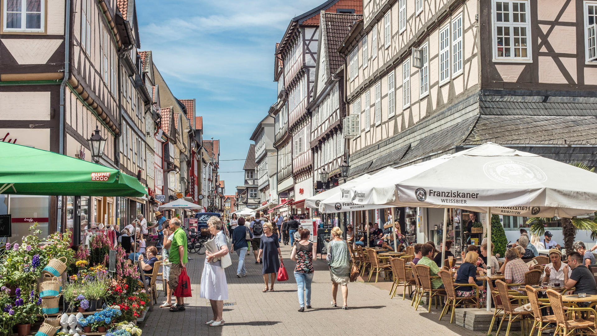 Auf einer gepflasterten Straße in der Innenstadt Wolfenbüttels befinden sich viele Personen. Einige davon stehen an Geschäften oder sitzen unter Sonnenschirmen. Links und rechts der Straße sind Fachwerkhäuser zu erkennen.