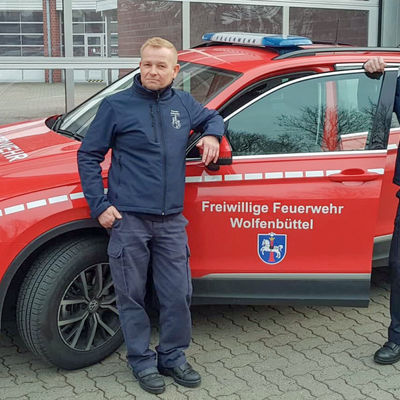 Vor einem roten Auto mit der Aufschrift "Freiwillige Feuerwehr Wolfenbüttel" stehen zwei Männer. Beide lehnen an jeweils einer Seite der offenen Fahrertür des Autos.